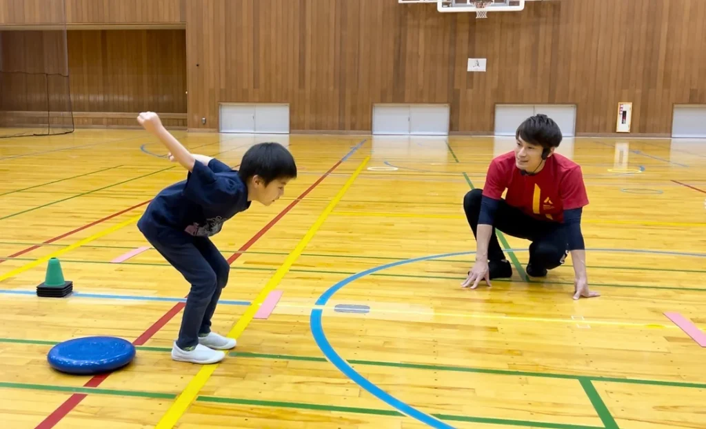 立ち幅跳びでジャンプをする直前の真剣な表情の小学生の子供と、その子を指導する大久保先生