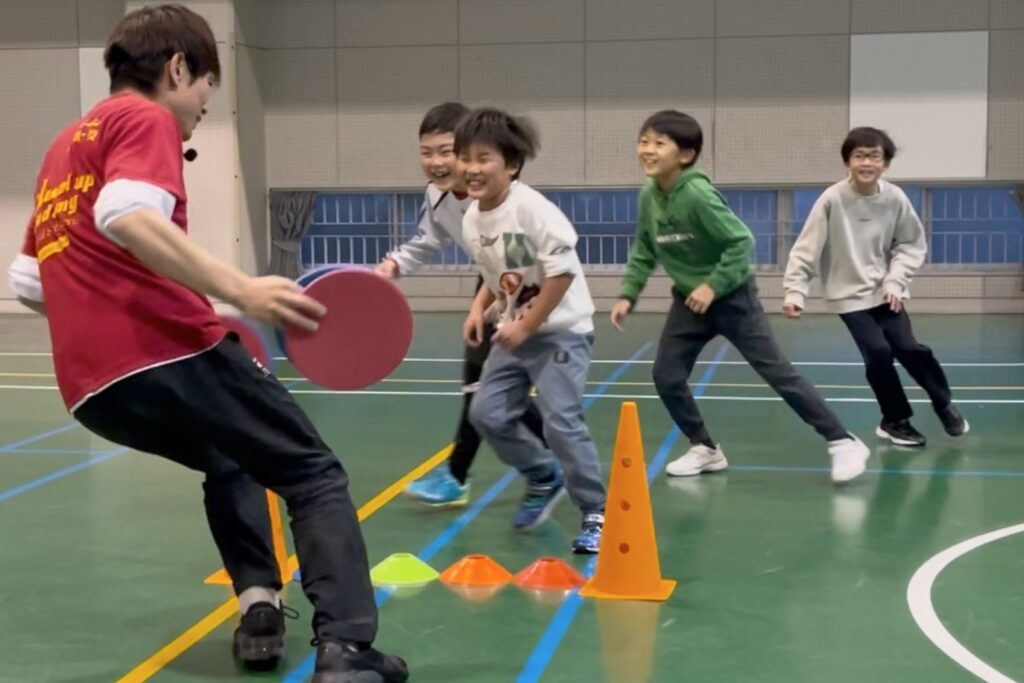 白山市教室にて、オリジナルの「かべ鬼ごっこ」をする万遍の笑顔の小学生の子供四人と、追いかける大久保先生